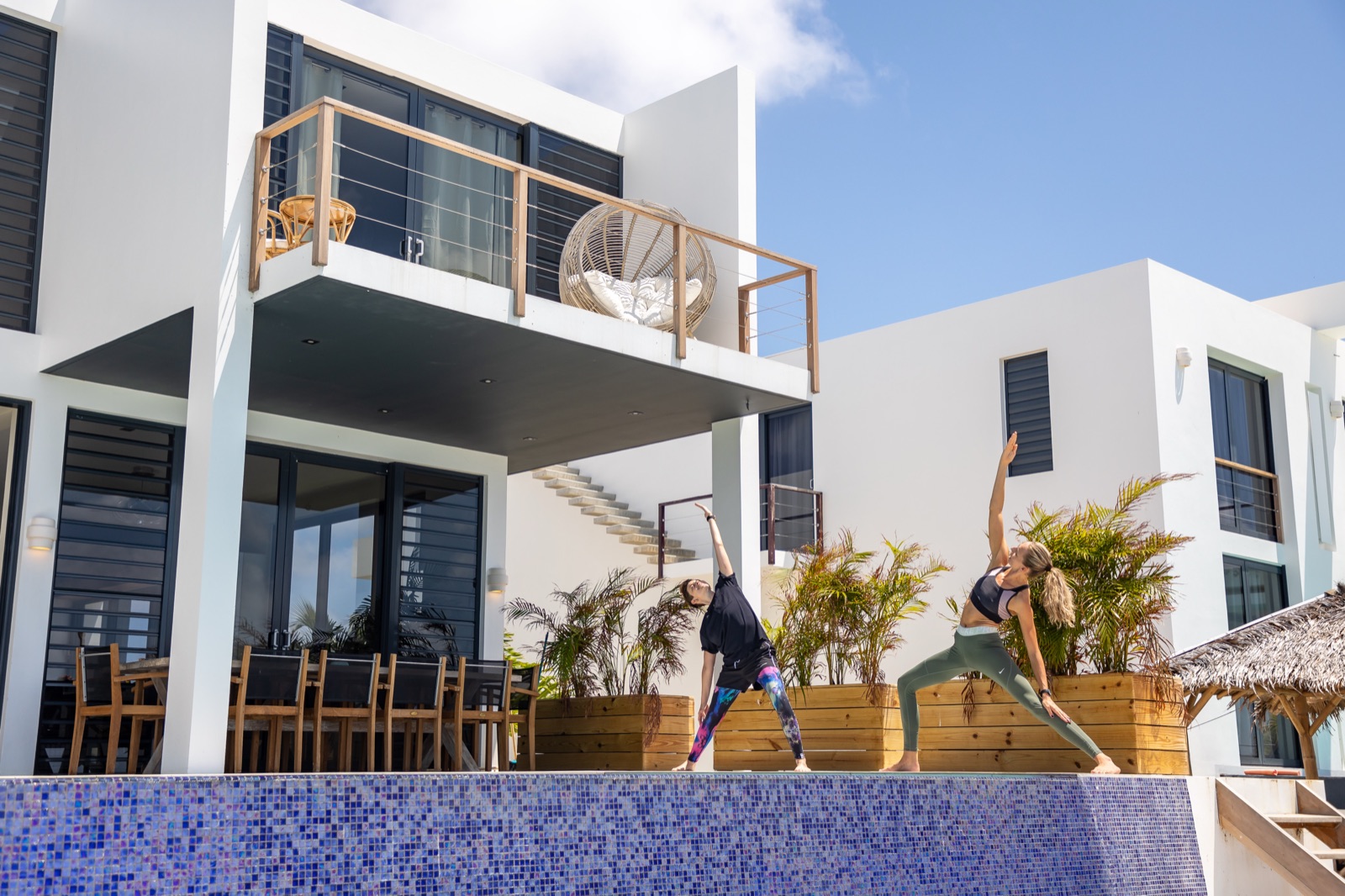 Two women practicing yoga by the private pool of a modern luxury Bonaire villa with white architecture and a second-floor balcony.
